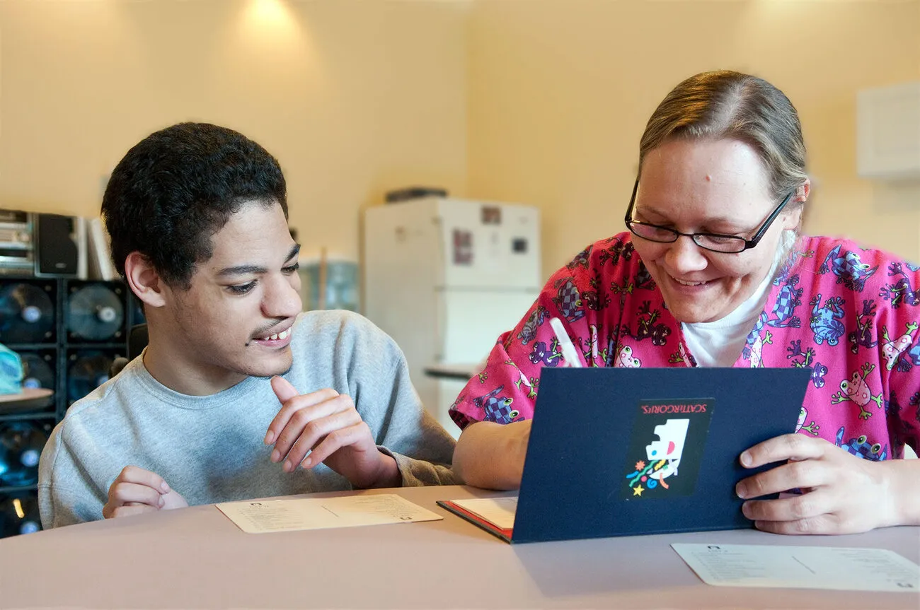 Caregiver in scrubs sharing a board game with a young adult client, both smiling at the table