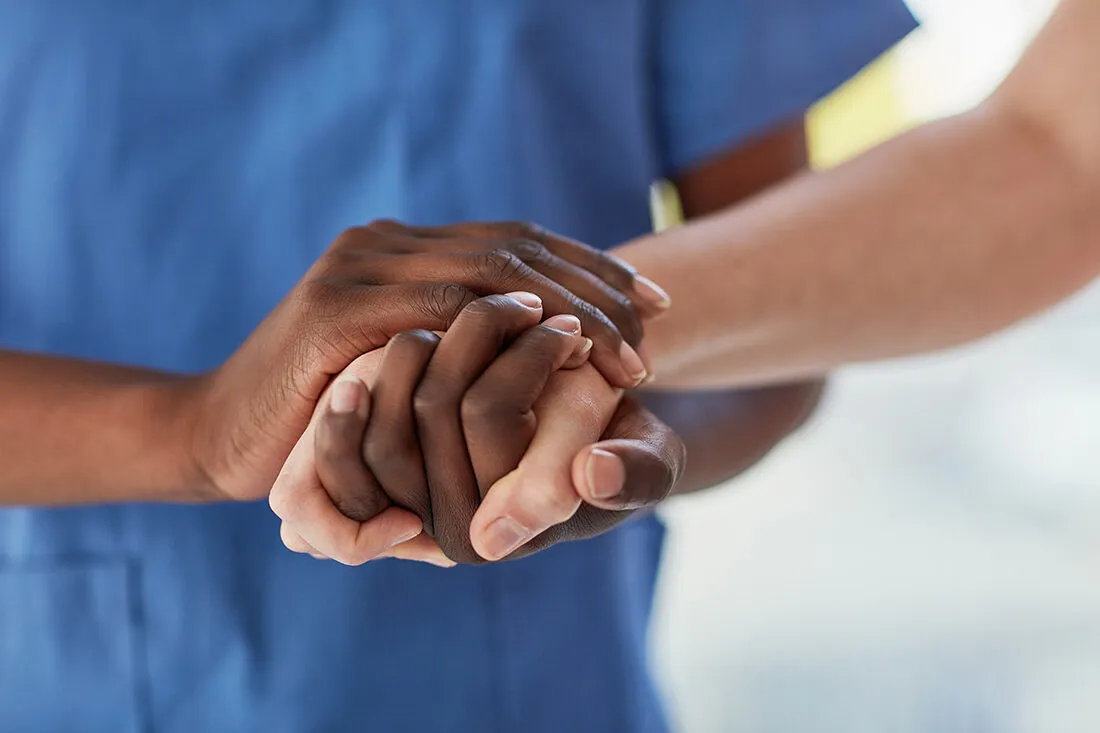 Black caregiver in blue scrubs holding the hands of an elderly white client — a moment of care