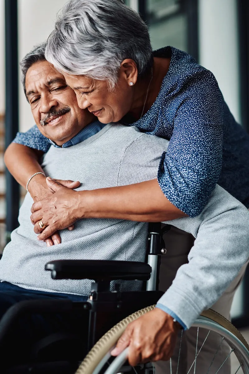 Older Hispanic woman tenderly embracing her husband from behind as he sits in a wheelchair
