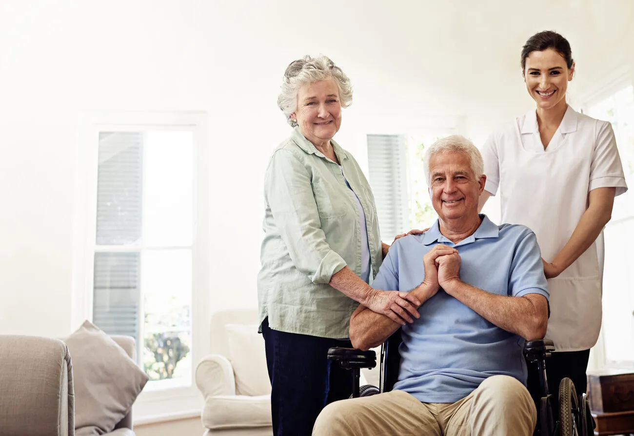 Senior couple with caregiver: husband in wheelchair, wife and caregiver both smiling in bright home