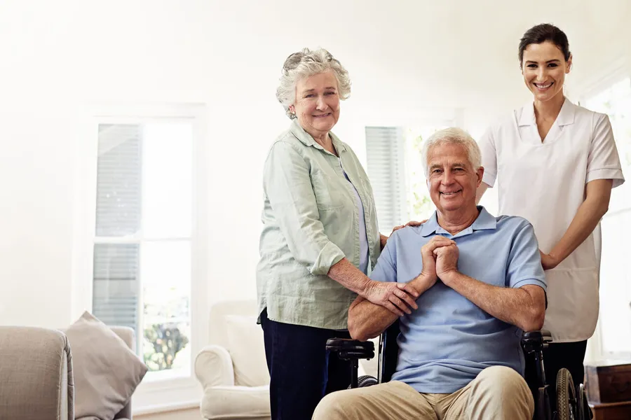 Senior couple with caregiver: husband in wheelchair, wife and caregiver both smiling in bright home