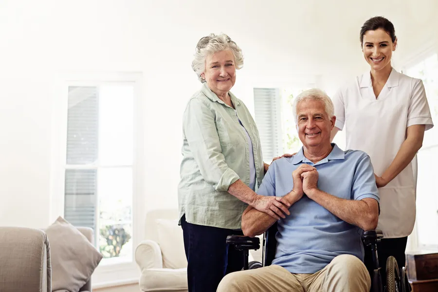 Senior couple with caregiver: husband in wheelchair, wife and caregiver both smiling in bright home