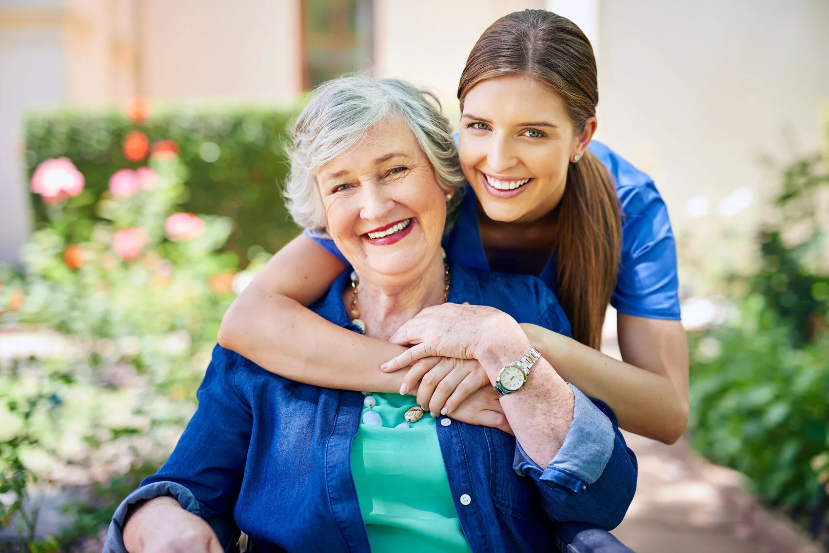 Young caregiver in blue shirt wrapping arms around smiling senior woman seated in a flower garden