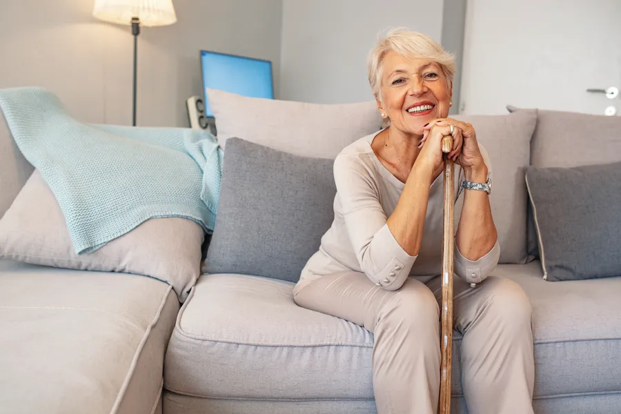 Active senior woman with short white hair sitting confidently on sofa, smiling with cane in hand