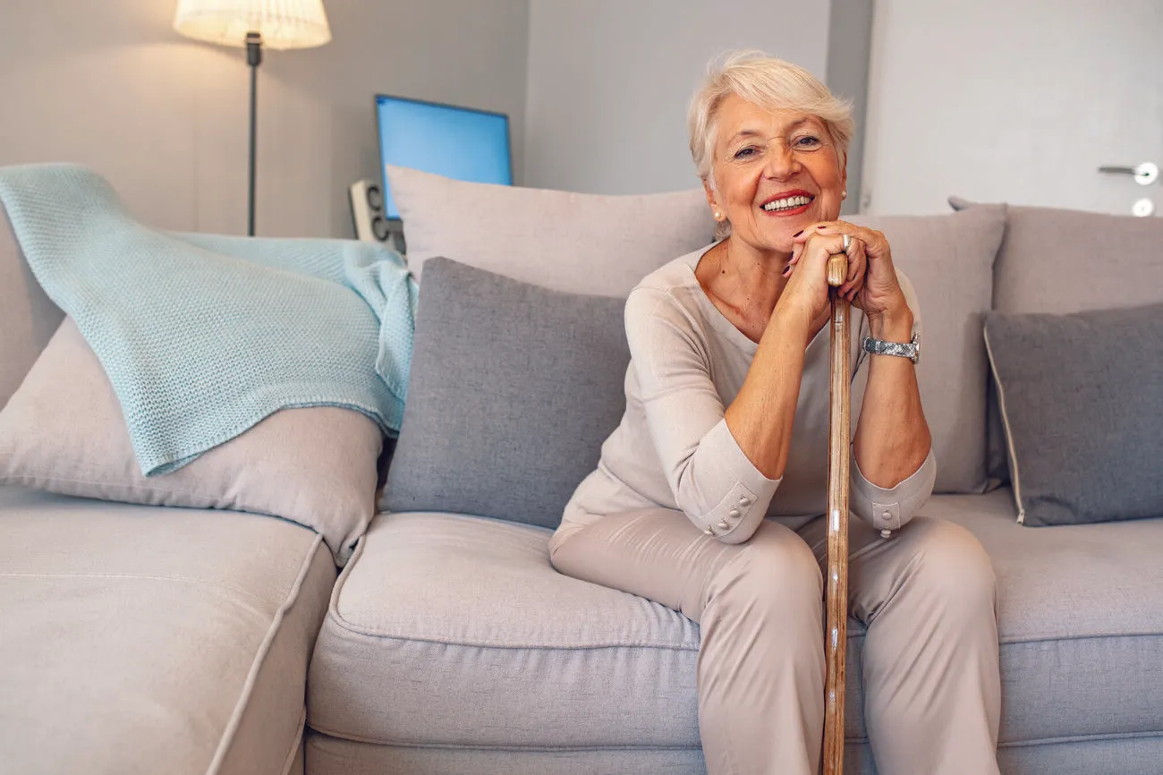 Active senior woman with short white hair sitting confidently on a sofa at home, smiling with a cane in hand
