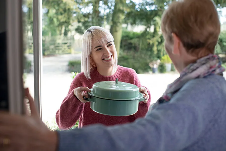 Caregiver handing a warm meal container to a smiling senior woman at her door