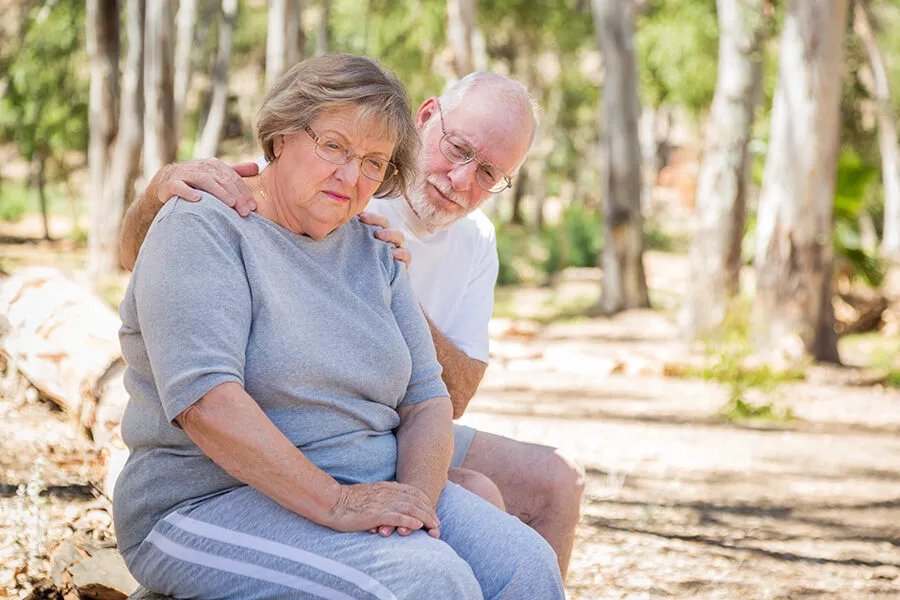 Older couple sitting together on a log outdoors among trees, husband gently supporting his wife