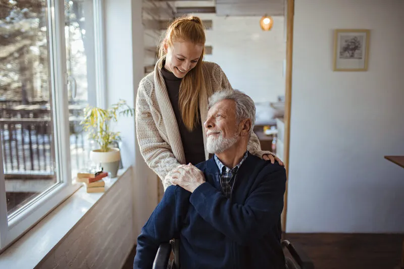 Daughter smiling warmly as she grasps the hand of her father in a wheelchair near a winter window
