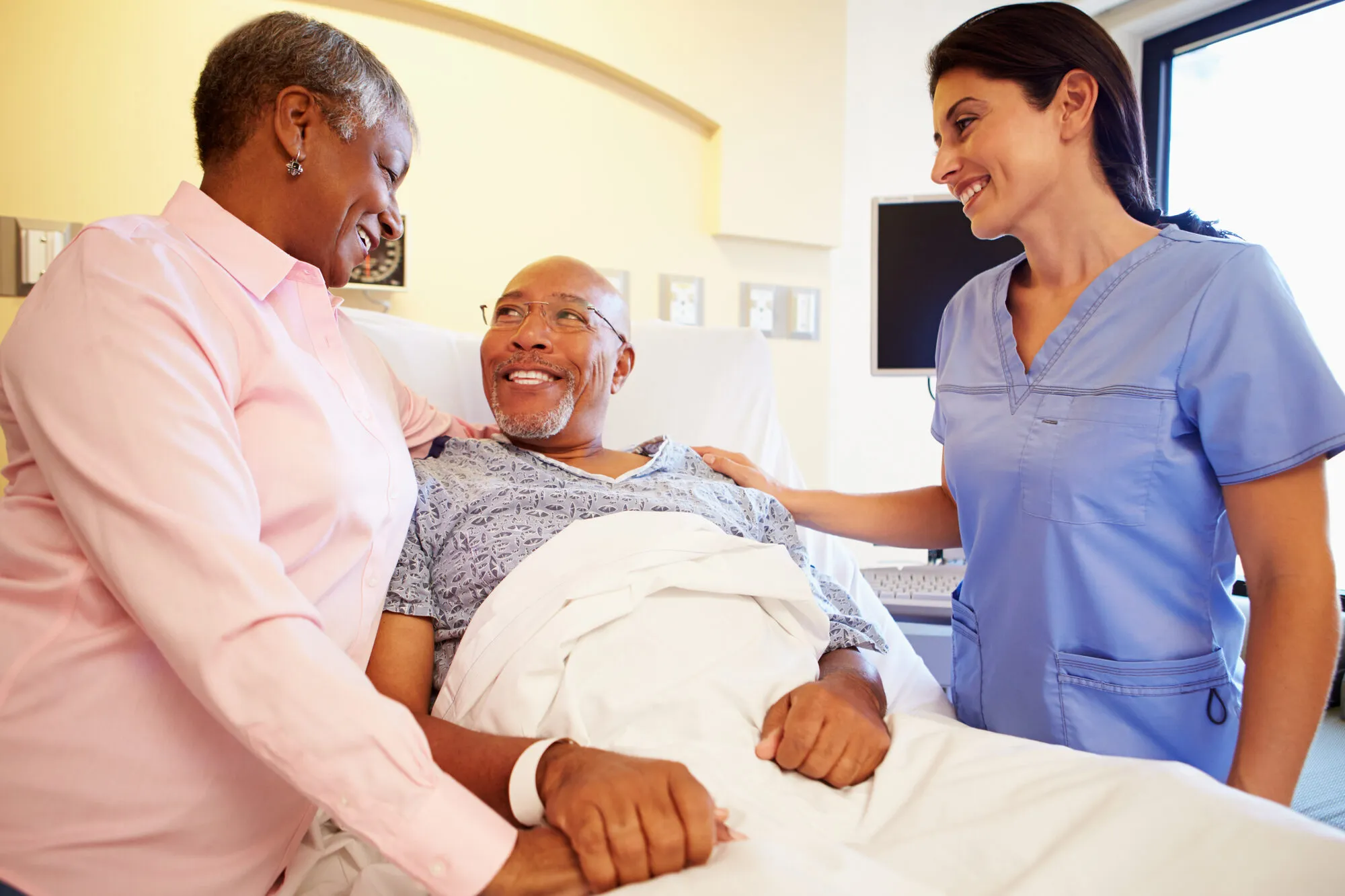 A smiling senior man recovering in a hospital bed with his wife and nurse at his side, beginning the transition to recovery at home