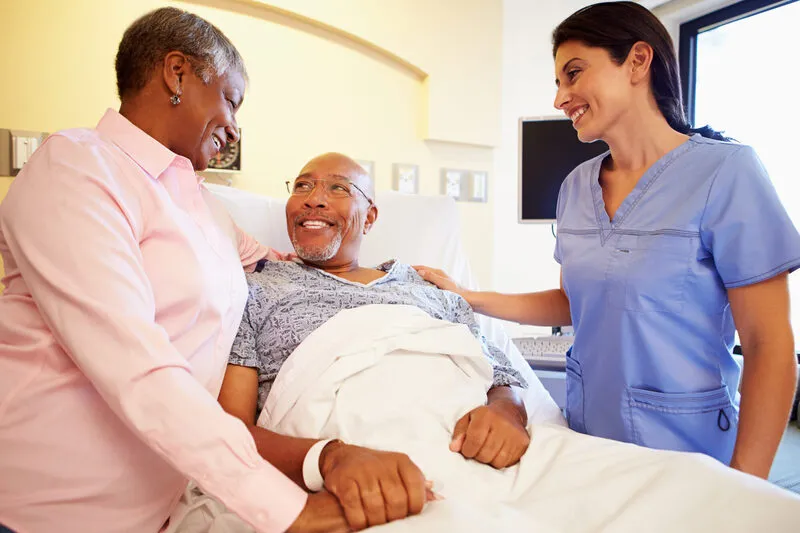 Black senior patient in hospital bed smiling, wife by his side, nurse reassuringly touching his shoulder