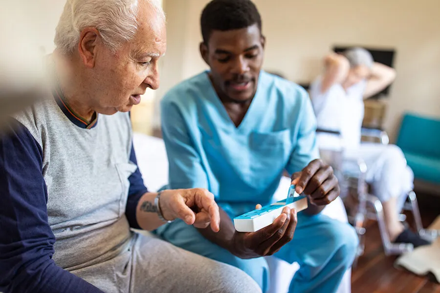 Young Black male caregiver in light blue scrubs helping an older white man review his weekly pill organizer