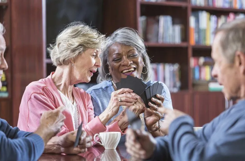 Two older women laughing while playing cards together over coffee in a book-lined room