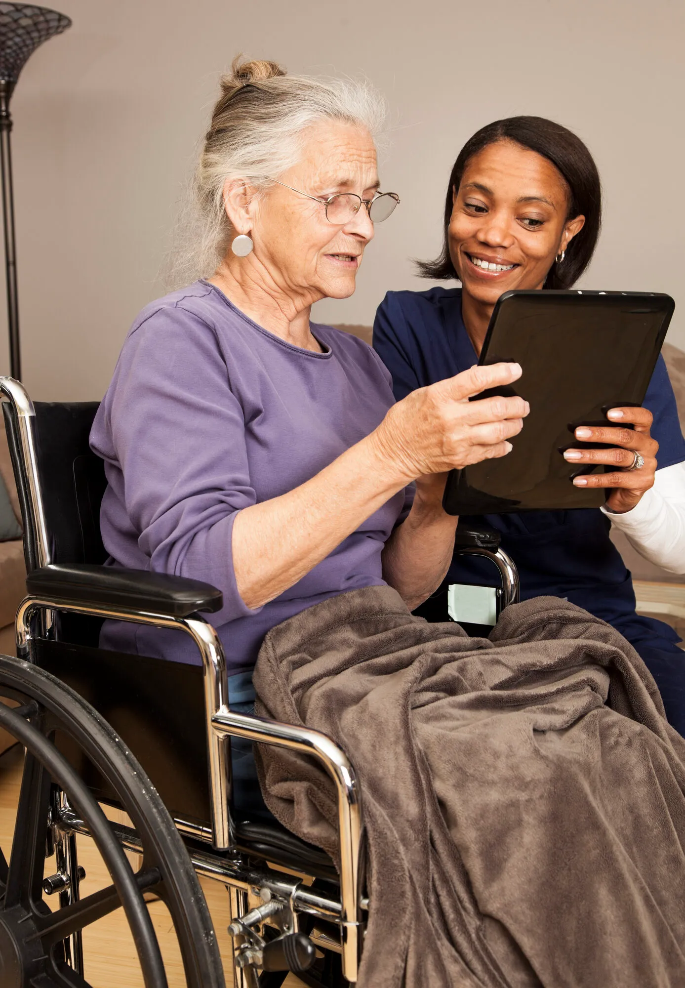 Black female caregiver showing a tablet screen to an elderly white woman in a wheelchair at home
