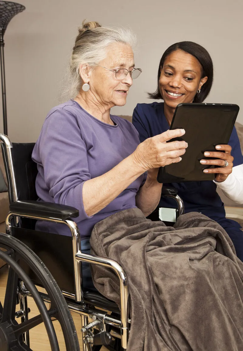 Black female caregiver showing a tablet screen to an elderly white woman in a wheelchair at home