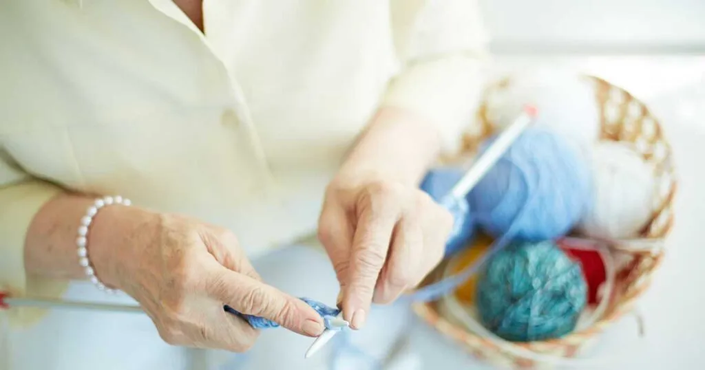 Close-up of senior woman's hands knitting with blue yarn next to a colorful basket of wool balls