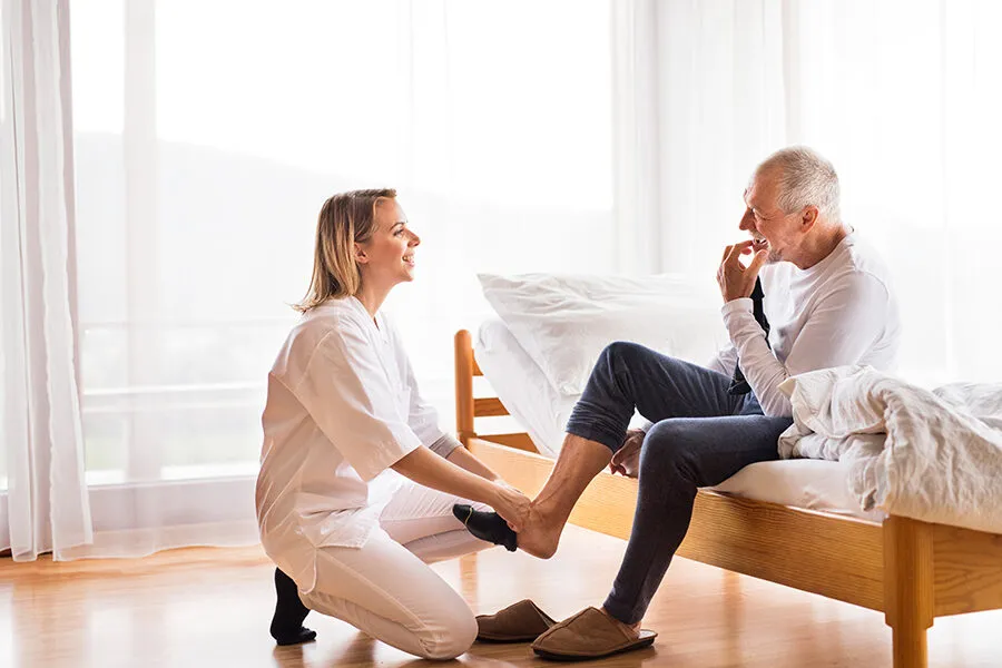 A SYNERGY HomeCare caregiver kneeling to help a smiling senior man put on socks and steady himself at the edge of his bed
