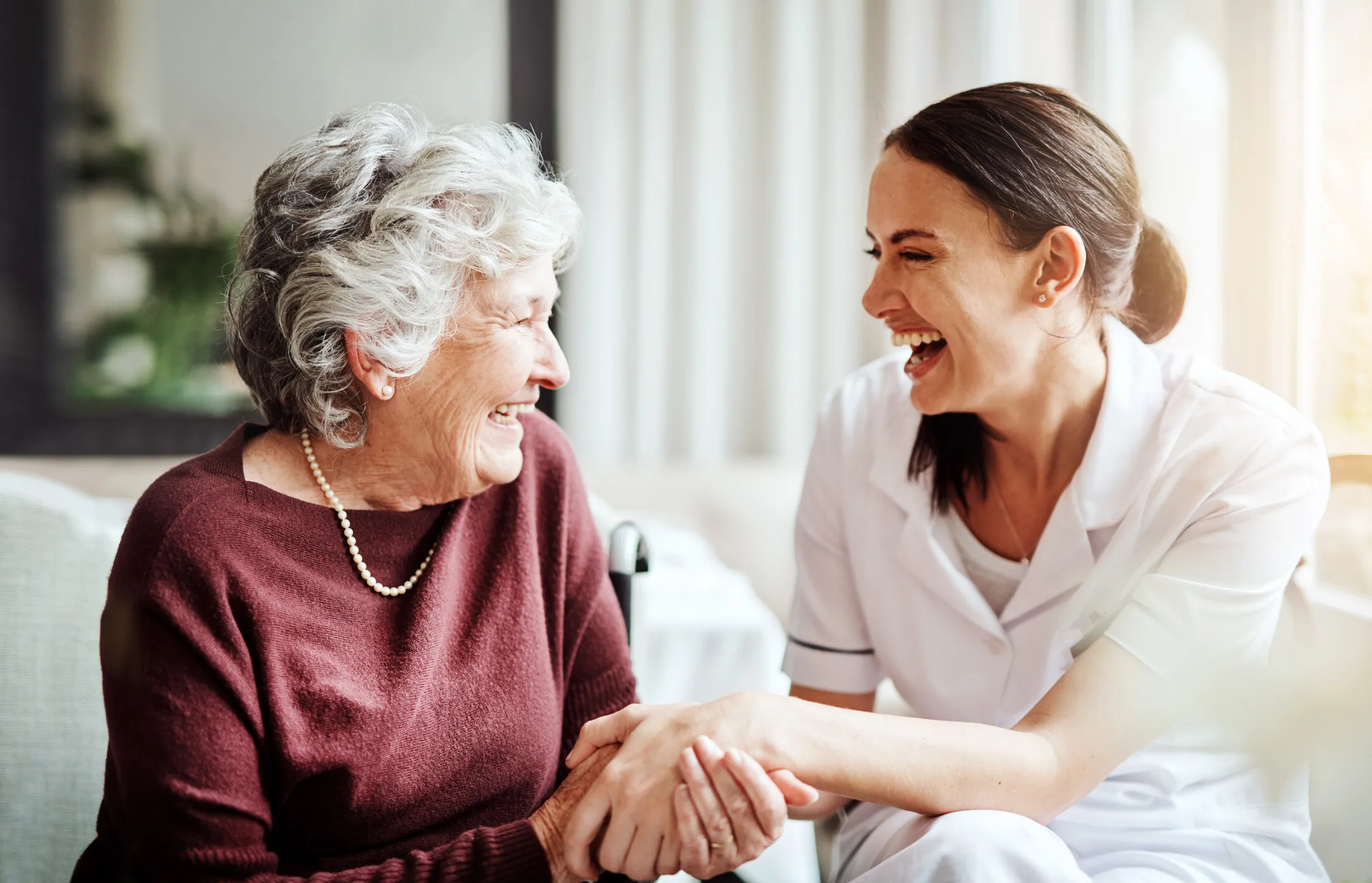 A SYNERGY HomeCare caregiver and a smiling senior woman laughing together and holding hands at home