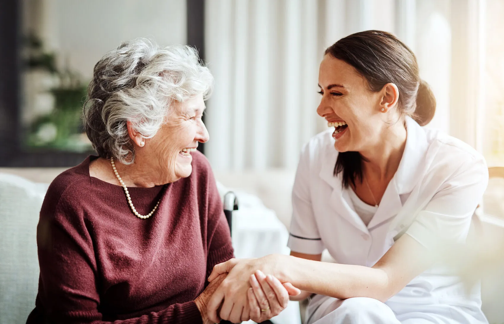 A SYNERGY HomeCare caregiver and a smiling senior woman laughing together and holding hands at home