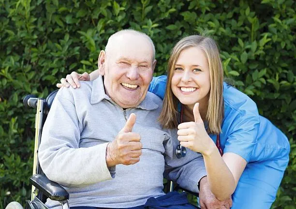 A SYNERGY HomeCare respite caregiver and a smiling senior man in a wheelchair giving a cheerful thumbs-up together outdoors